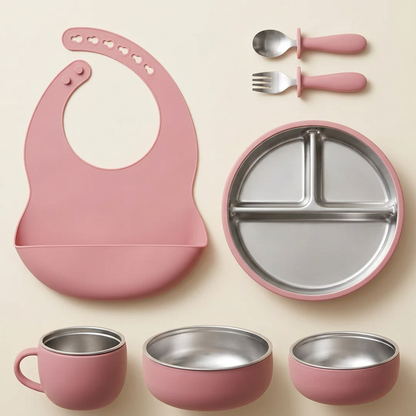 Single powder-rose seven-piece mealtime set with silicone bib, divided steel plate, cup, two bowls, fork and spoon on warm ivory